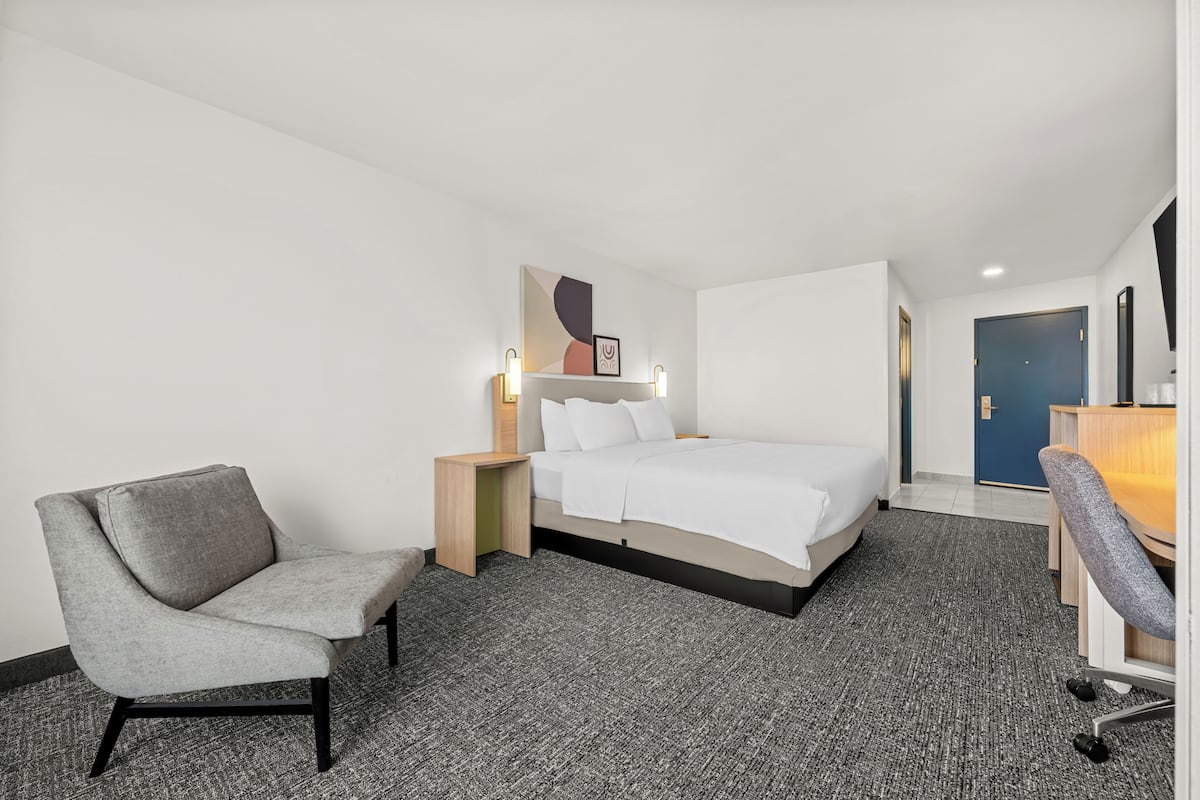 A hotel room with a bed, two chairs, a desk, and a wall-mounted artwork.