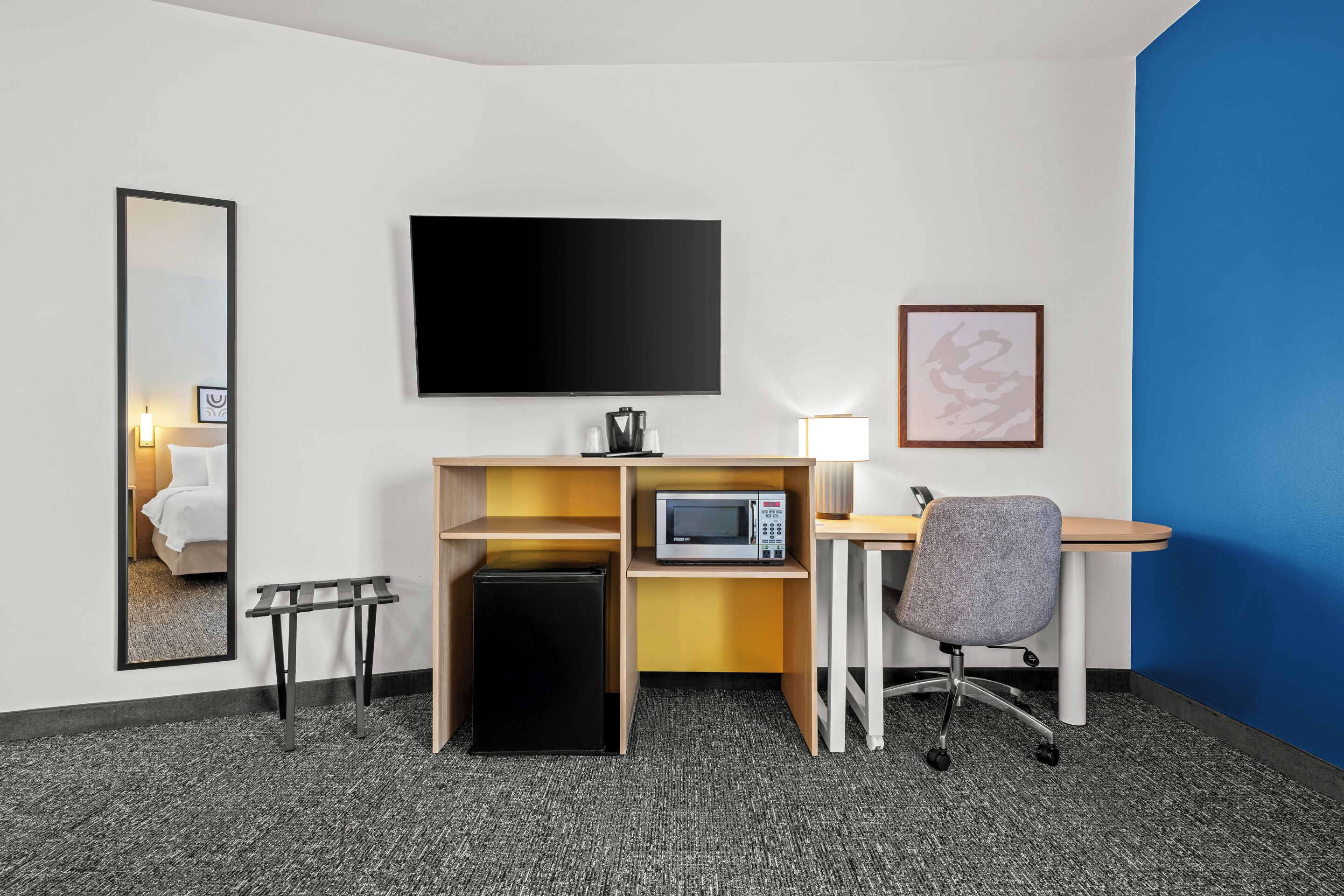 A hotel room with a bed, a desk with a computer, a microwave, a small refrigerator, a chair, a flat-screen TV, a bedside table, and a mirror.
