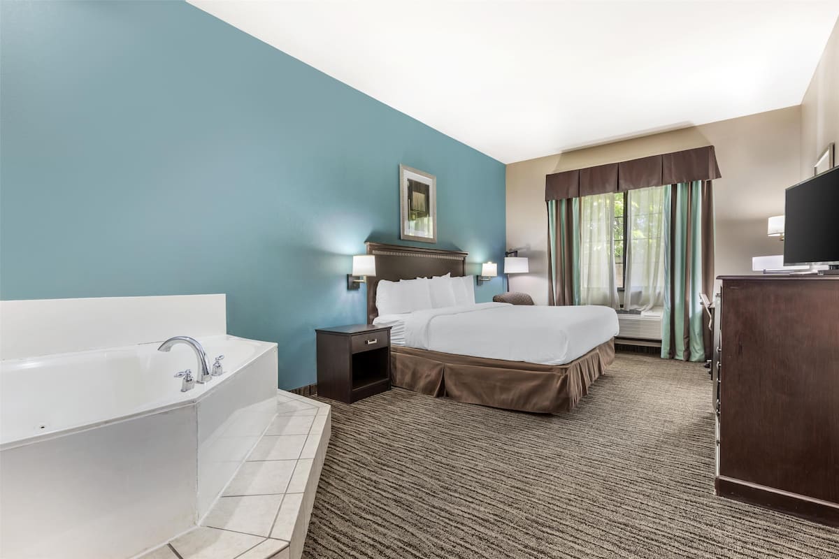 A hotel room with a large bed, a separate bathroom with a bathtub, a desk with a television, and a window with curtains.