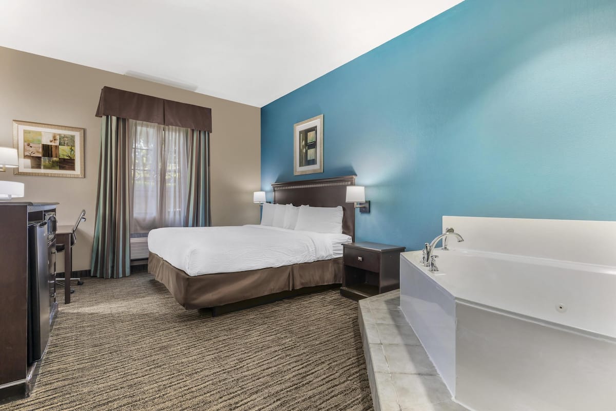 A hotel room with a bed, a desk, a chair, a dresser, a large window with curtains, a wall-mounted picture, a nightstand with a lamp, and a bathtub.