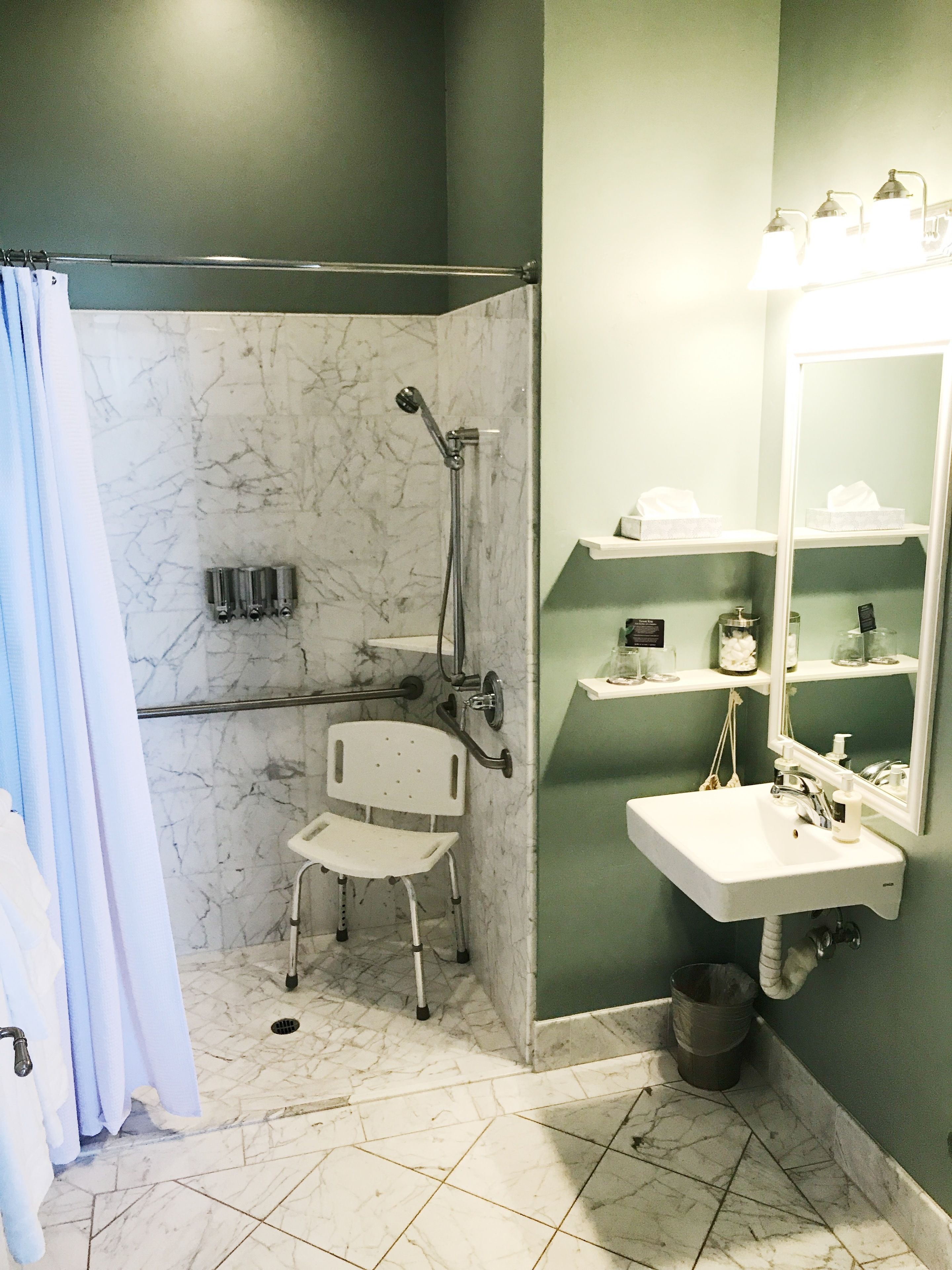 A bathroom with a shower, a white chair, a sink, and a mirror.