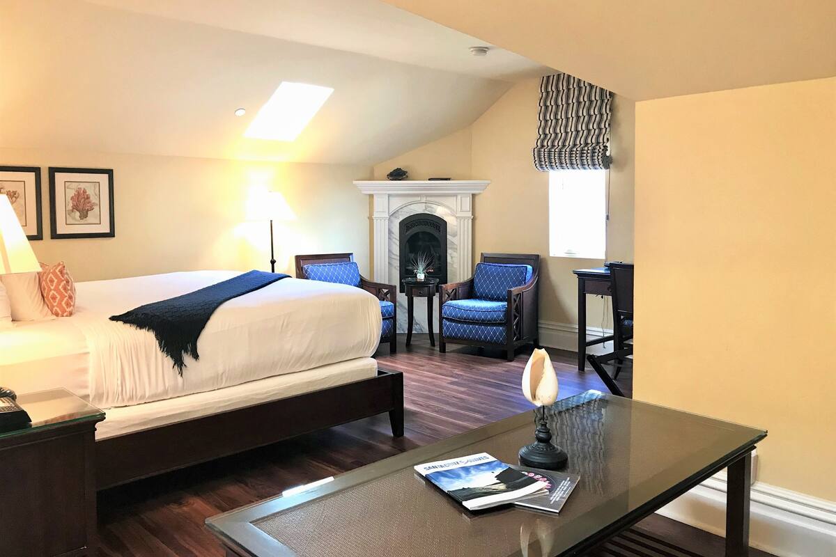 A hotel room with a bed, a fireplace, a chair, a table with a lamp, and framed pictures on the wall.