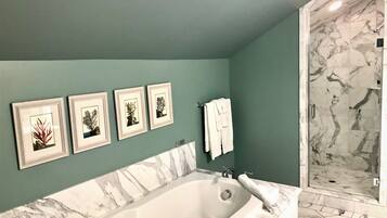 A bathroom with a large bathtub, a shower with marble walls, and four framed botanical prints on the wall.
