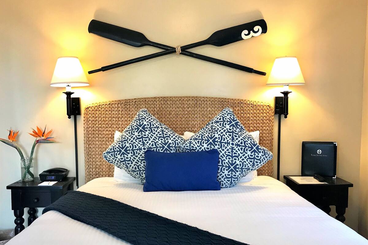 A hotel room with a bed, two bedside tables, a wall-mounted lamp, a decorative wall piece, and a vase with flowers.