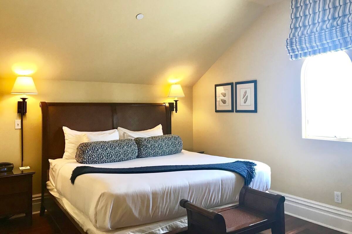 A bedroom with a large bed, two bedside lamps, a nightstand, a chair, and a window with curtains.