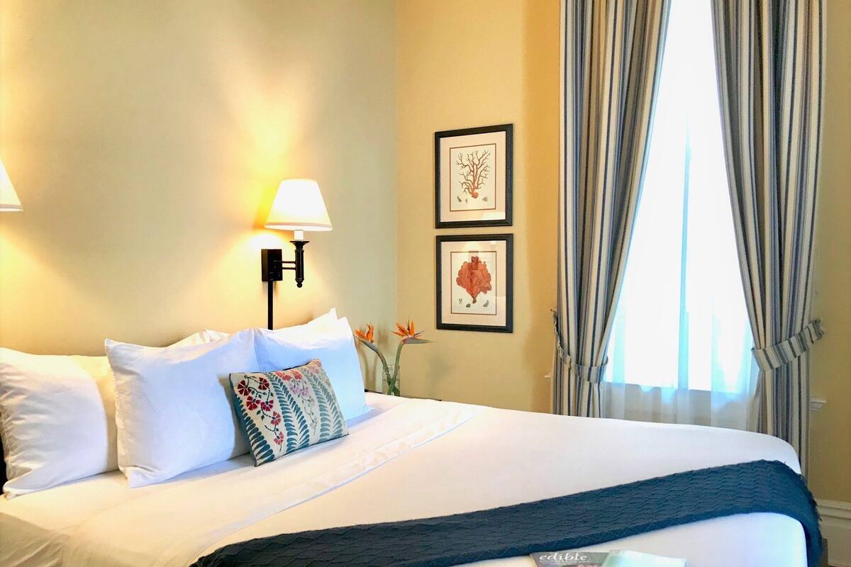 A hotel room with a neatly made bed, a lamp, framed botanical prints, and a window with curtains.