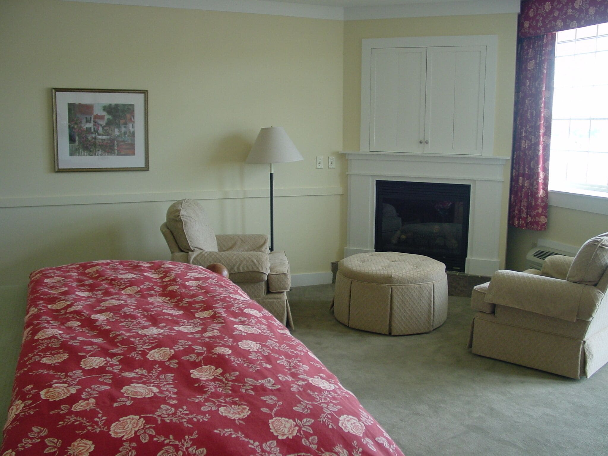 A bedroom with a bed, two armchairs, a side table, a lamp, a fireplace, and a framed picture on the wall.