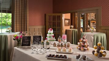 A dessert table with various cakes, cupcakes, and chocolates, set in a room with patterned wallpaper and large windows.