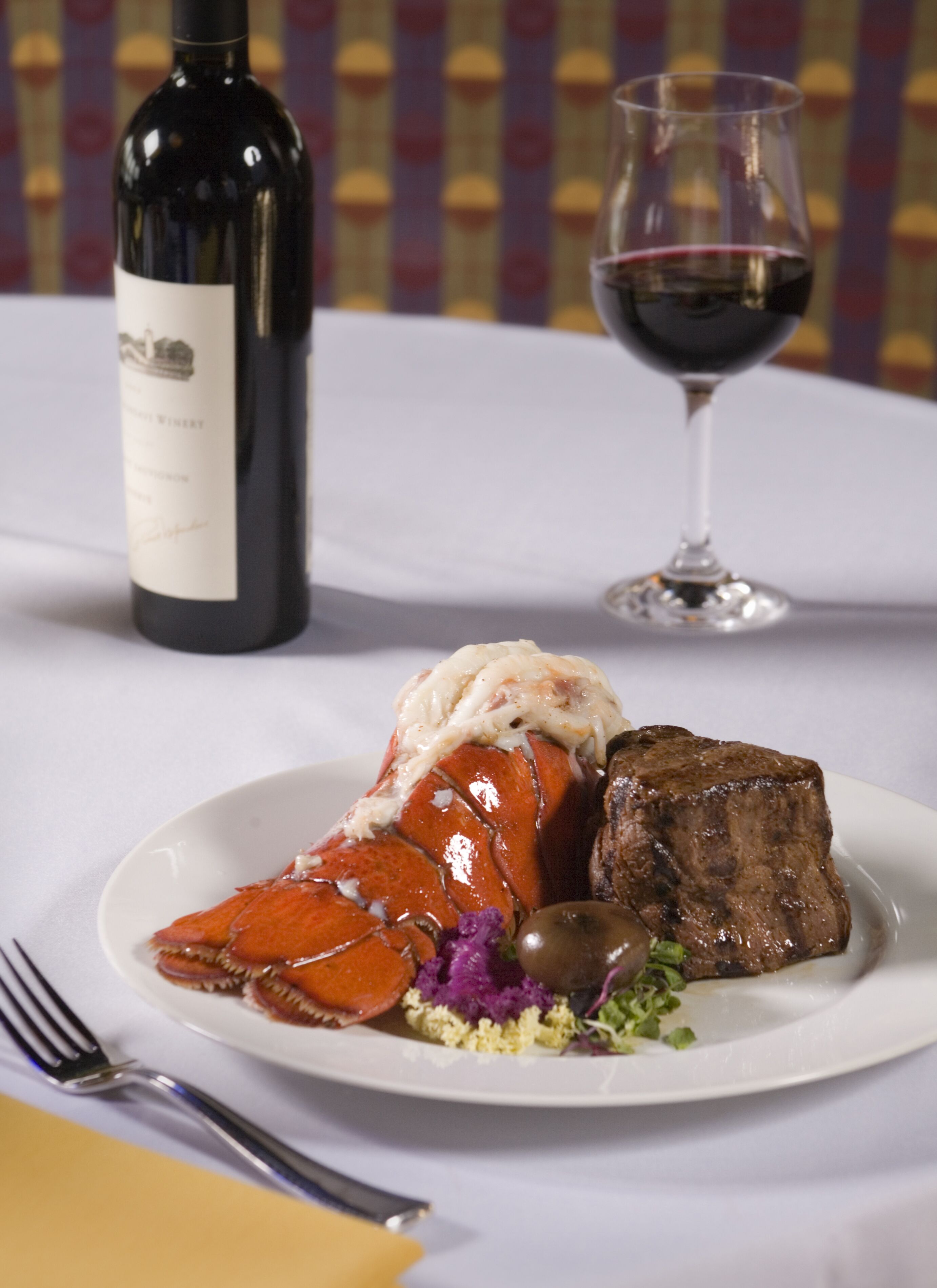 A plate with lobster, steak, and vegetables, a wine bottle, and a wine glass on a table.