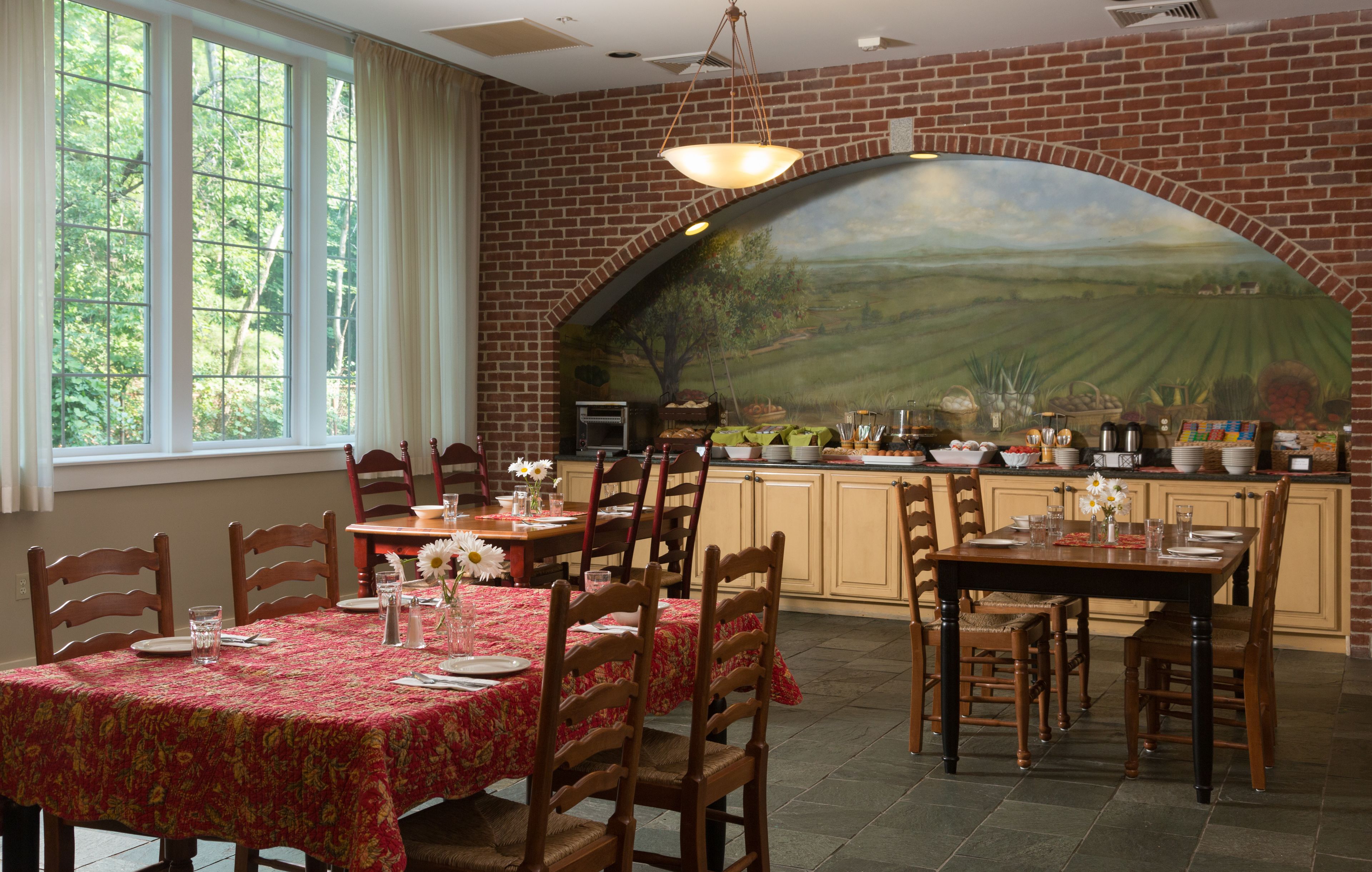 A dining area with a brick wall mural, wooden chairs, a table with a red tablecloth, and a kitchen area with a brick arch.