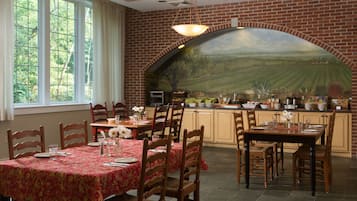 Un espace repas agrémenté d’une fresque murale en briques, de chaises en bois, d’une table recouverte d’une nappe rouge et d’un coin cuisine avec une arche en briques.