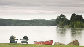 Two Adirondack chairs by a lake with a red boat and a small cabin in the background.