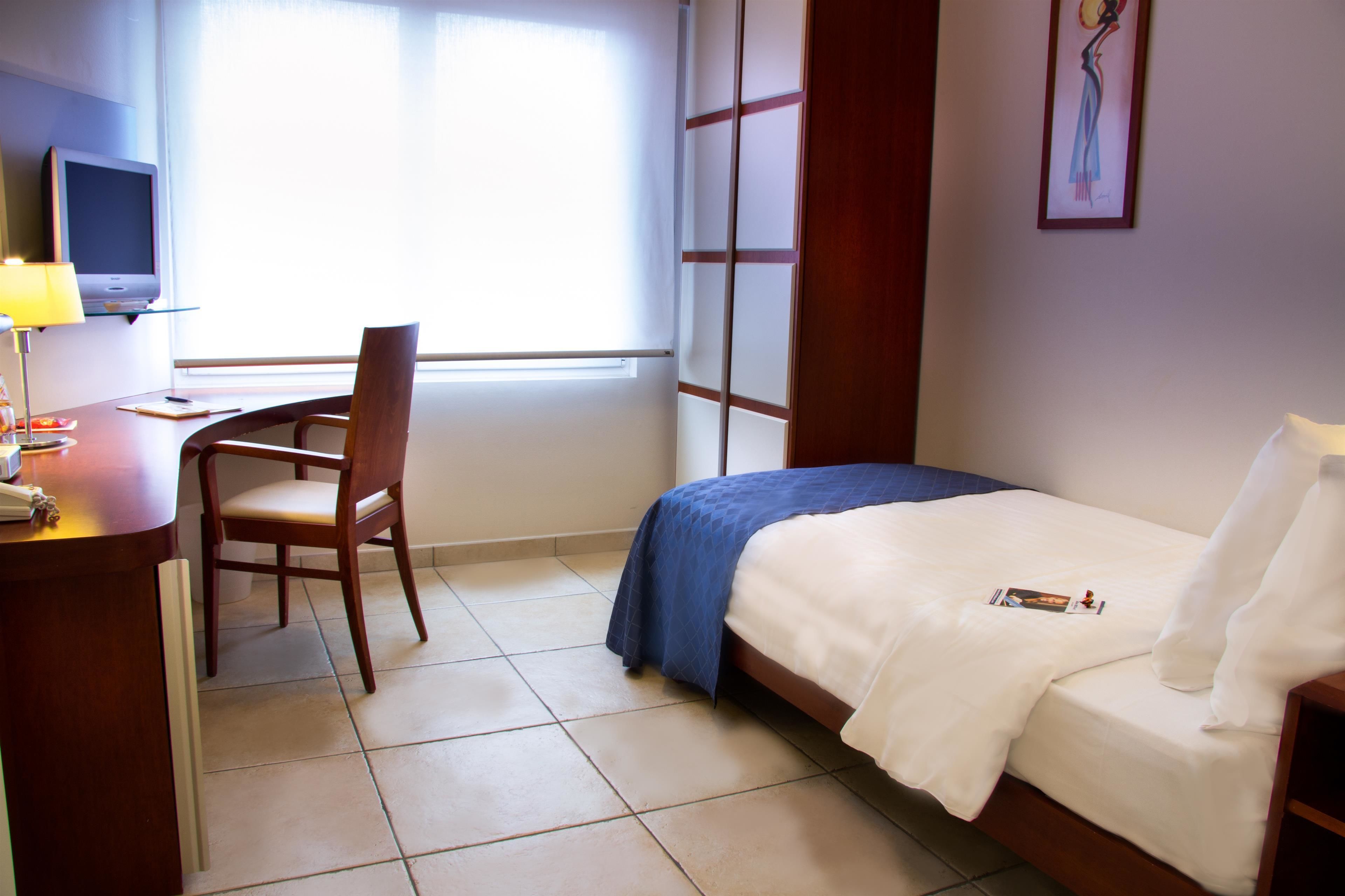 A hotel room with a bed, a desk with a lamp, a chair, a TV, and a window with blinds.