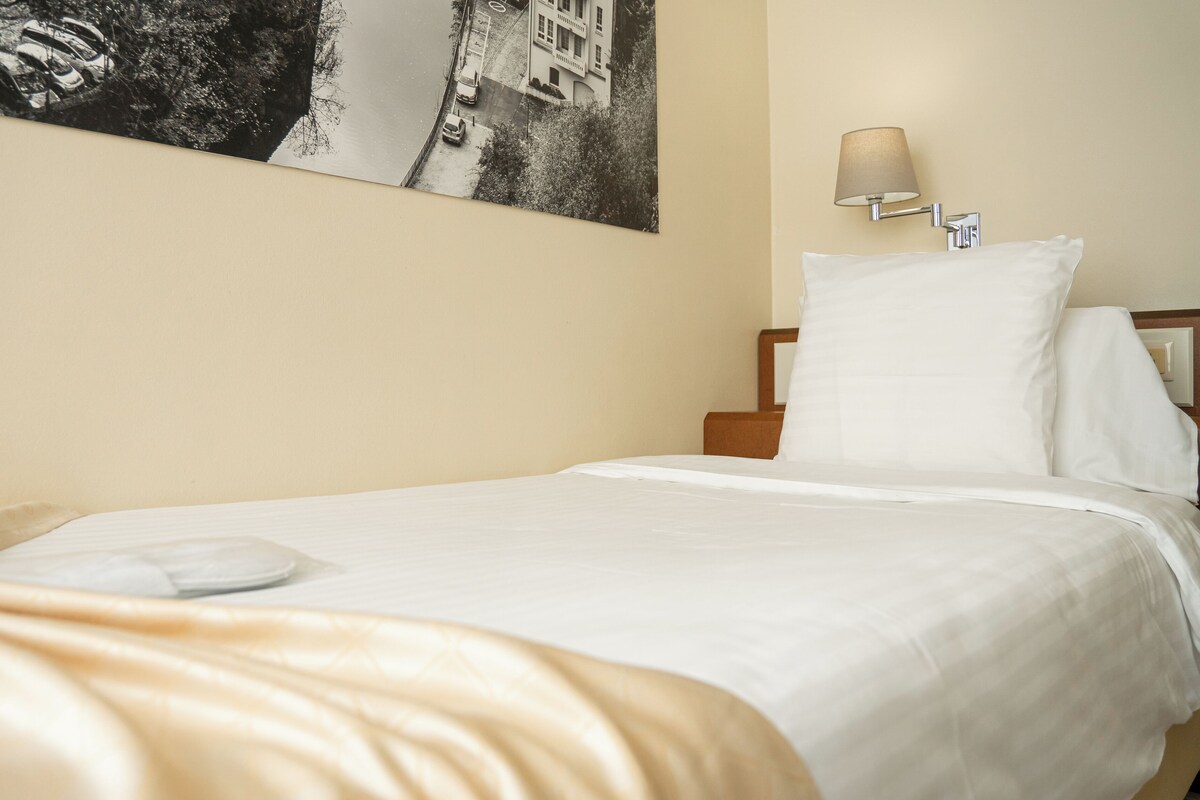 A neatly made bed with white linens, a wall-mounted lamp, and a framed picture above the bed.