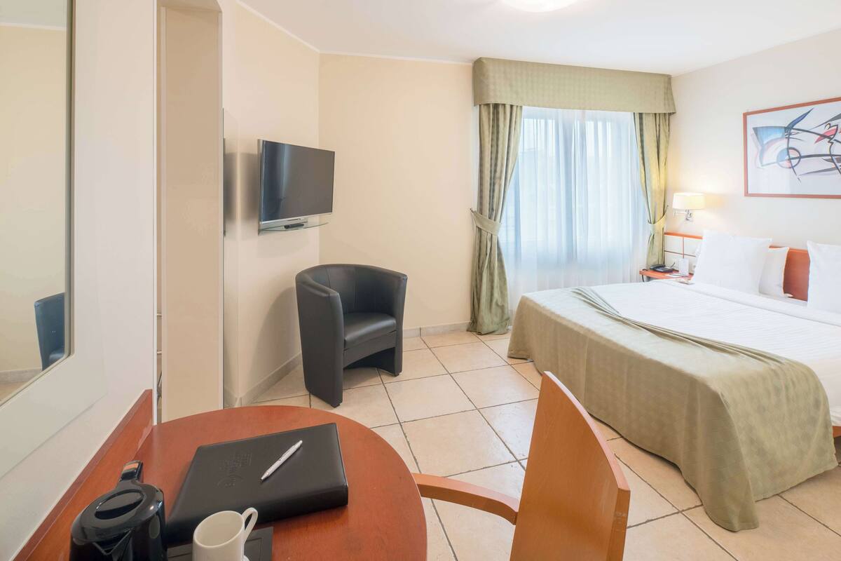 A hotel room with a bed, a desk with a laptop, a chair, a TV, and a window with curtains.