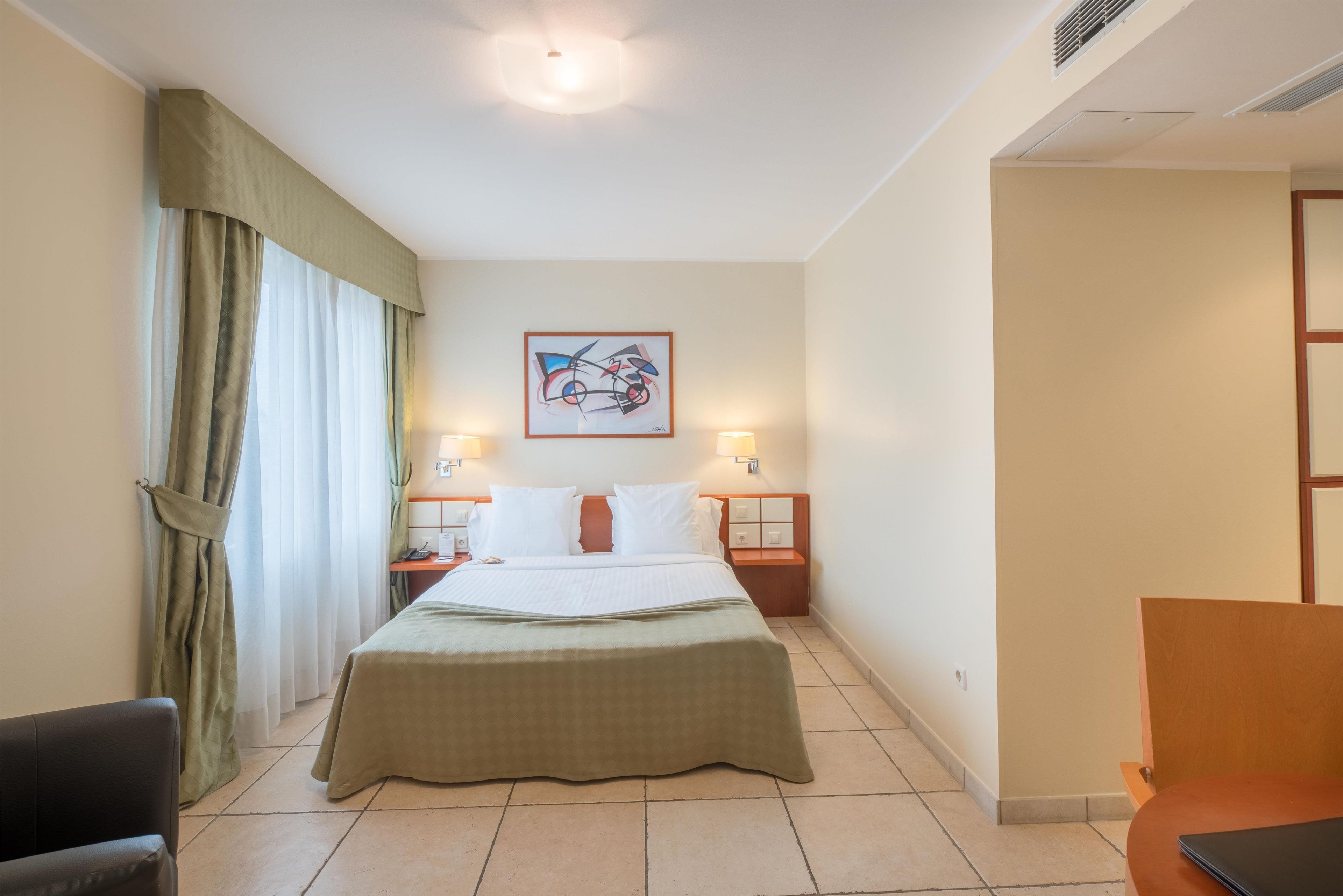Quarto de hotel com uma cama, duas mesas de cabeceira, uma escrivaninha, uma cadeira, uma janela com cortinas e um quadro na parede.