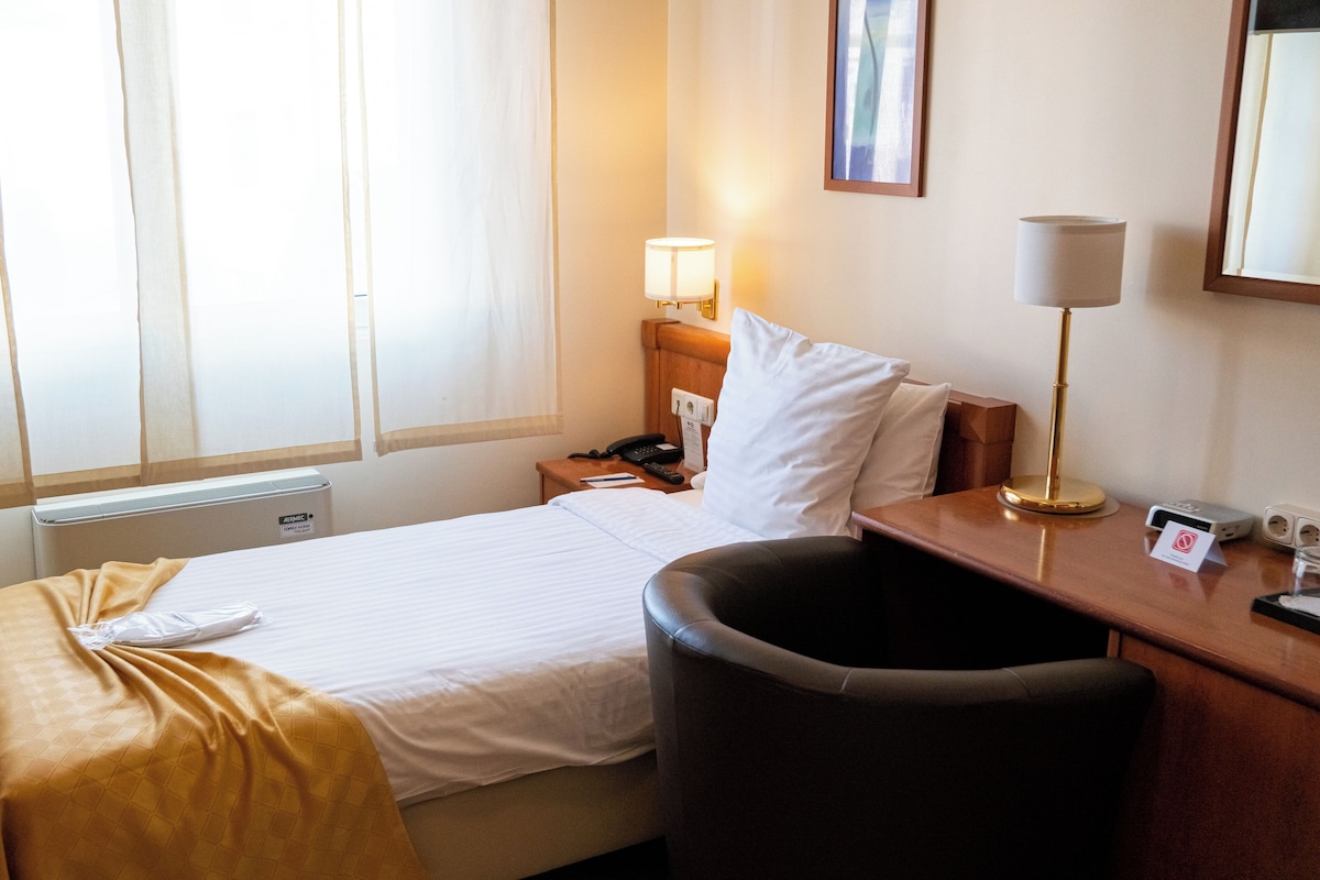 A hotel room with a bed, a desk, a chair, a lamp, a phone, and a window with curtains.