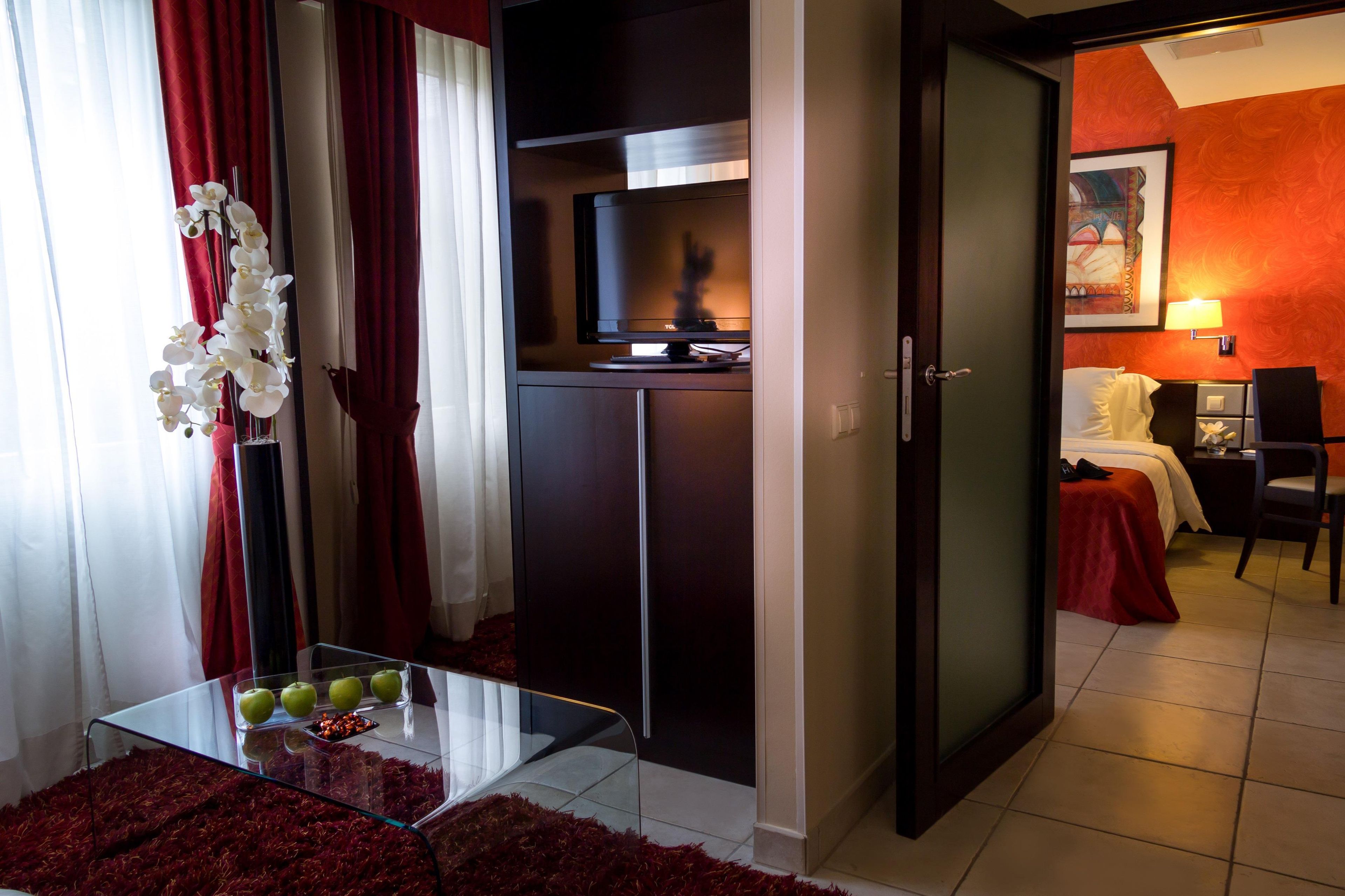 A hotel room with a glass table, red curtains, a TV, a bed, and a red carpet.