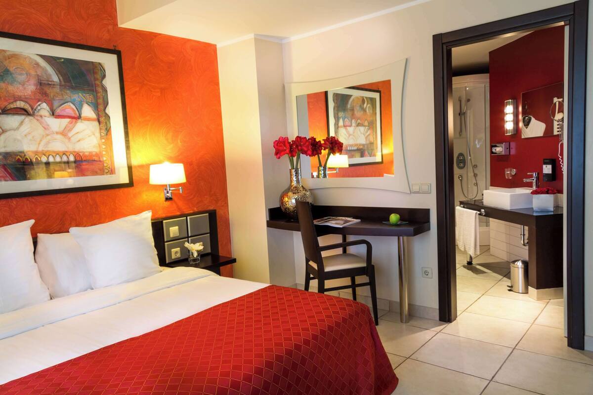 A hotel room with a bed, a desk, a chair, a vase with flowers, a painting on the wall, and a bathroom visible through an open door.