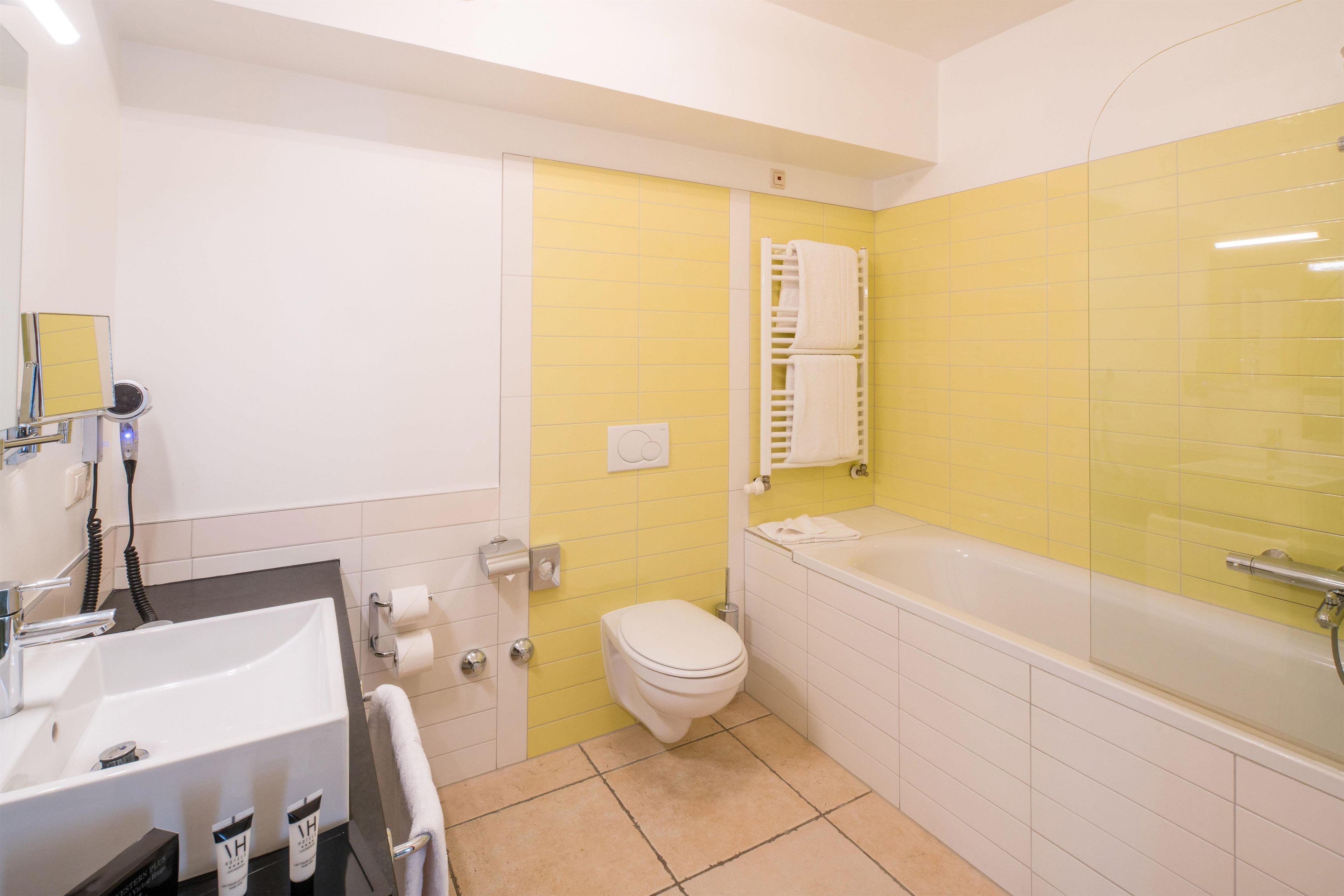 Une salle de bain avec un lavabo, des toilettes et une baignoire, tous de couleur blanche. On y trouve un porte-serviettes et un miroir au-dessus du lavabo.