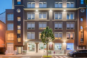 A multi-story hotel with a central entrance, a tree in the foreground, and a car parked on the street.
