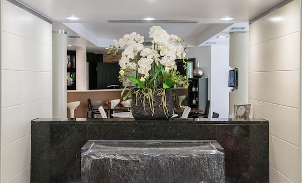 A reception desk with a black marble top, a vase of white orchids, and a small water feature in front. - Best Western Plus Grand Hotel Victor Hugo (Luxembourg City)