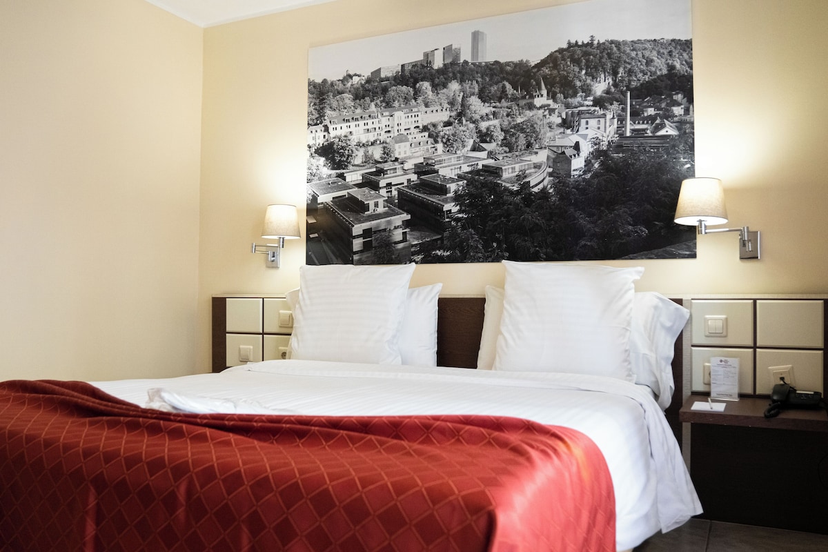 A hotel room with a bed, a large cityscape poster on the wall, two bedside lamps, and a nightstand with a phone and a notepad.