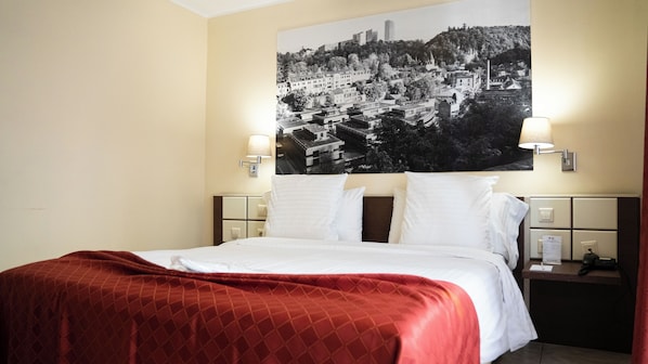 Una habitación de hotel con una cama, un cartel grande con vistas a la ciudad, dos lámparas de noche y una mesita de noche con un teléfono y un bloc de notas.