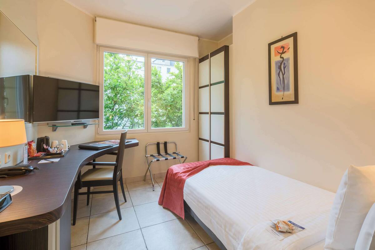 A hotel room with a bed, desk, chair, TV, and a window with a view of trees.