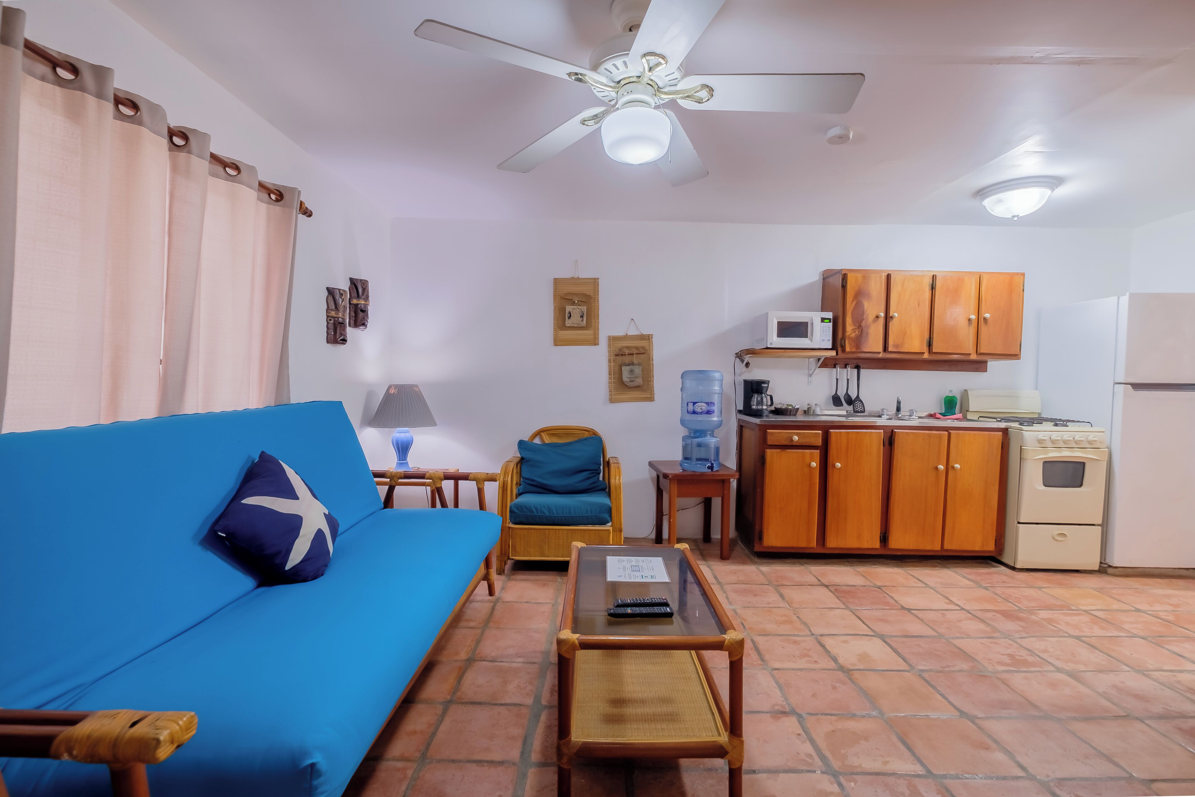 Um espaço compacto para viver, com um sofá azul, uma pequena mesa, uma cadeira, armários de madeira, uma kitchenette e um ventilador de teto.