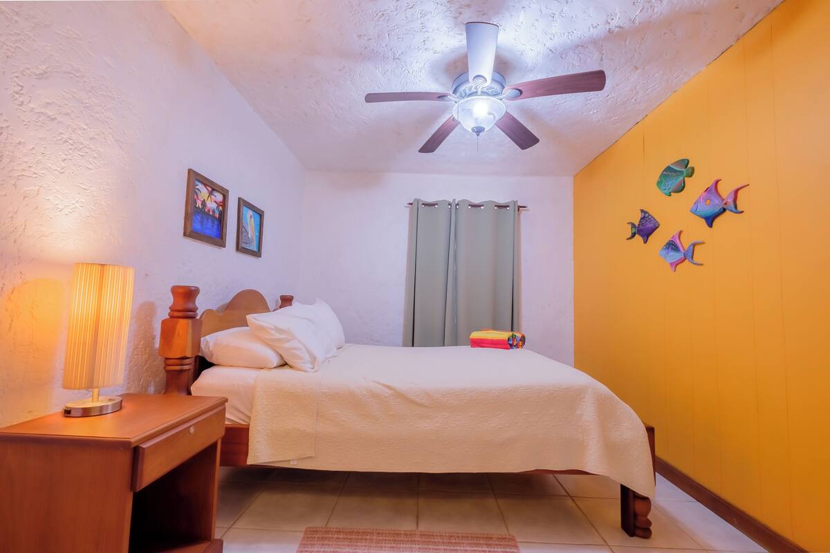 Un dormitorio con cama, mesita de noche, lámpara, ventilador de techo y decoración en la pared.