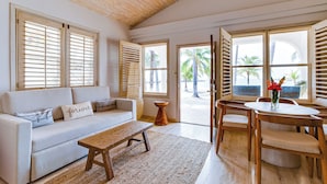 Amplia sala de estar con sofá blanco, mesa de centro de madera y una mesa de comedor con sillas. Amplias ventanas con contraventanas de madera ofrecen abundante luz natural y vistas a palmeras.