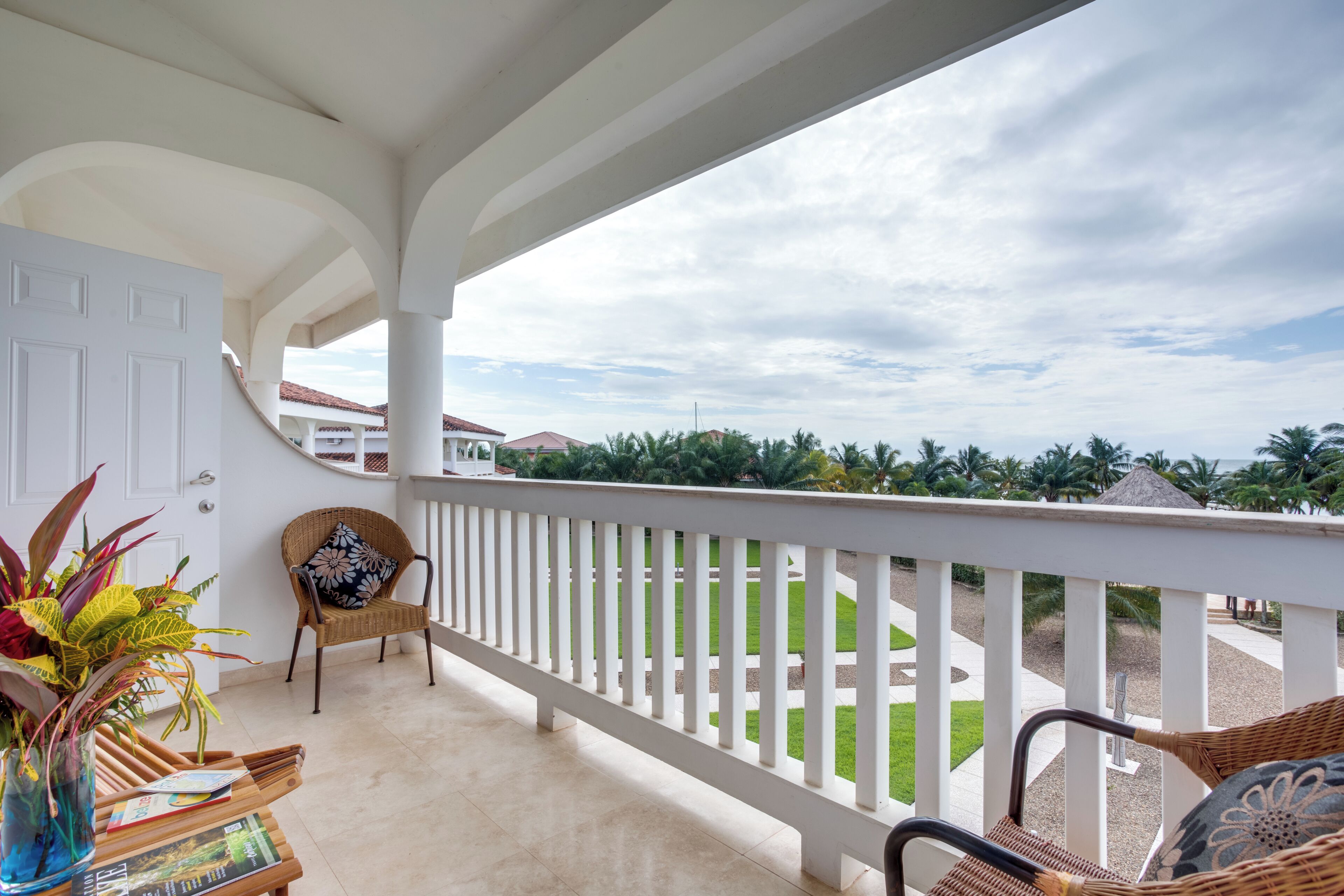 A balcony with a white railing, a wicker chair, a potted plant, and a view of a residential area with houses and palm trees.