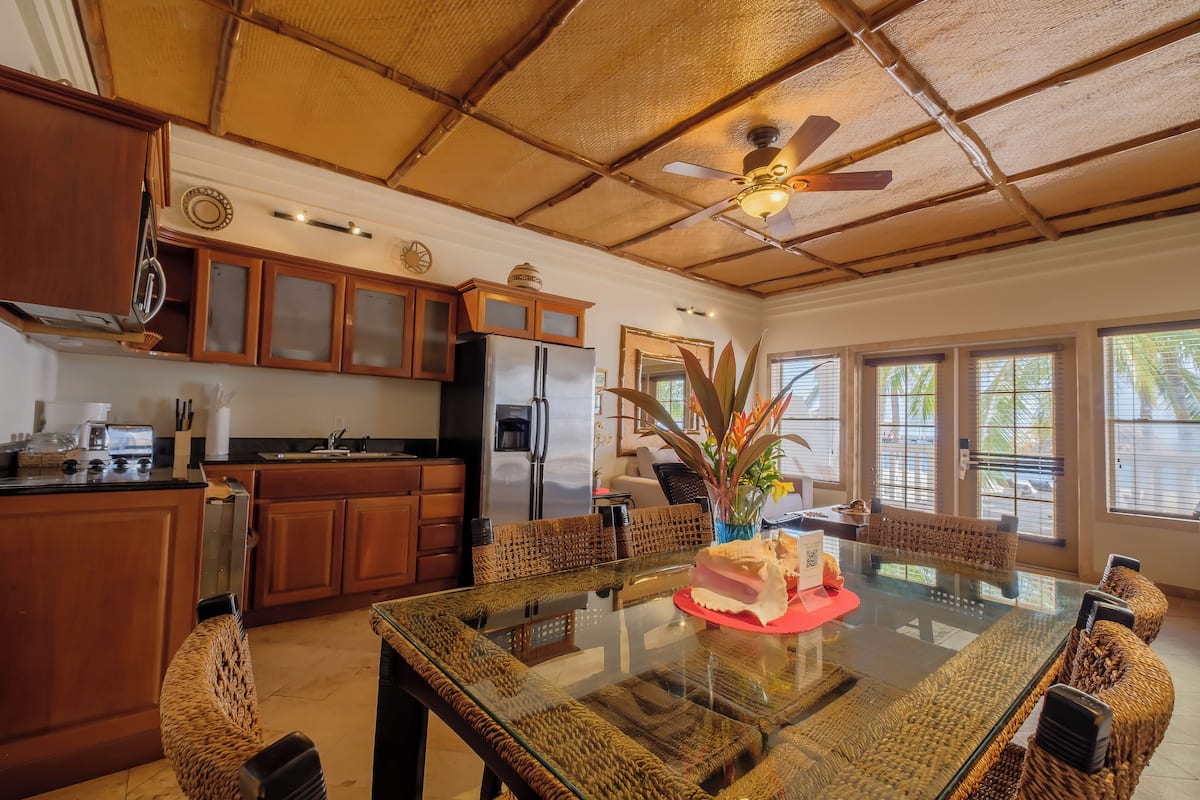 A spacious kitchen with wooden cabinets, a large island, and a dining area with a glass table and woven chairs.