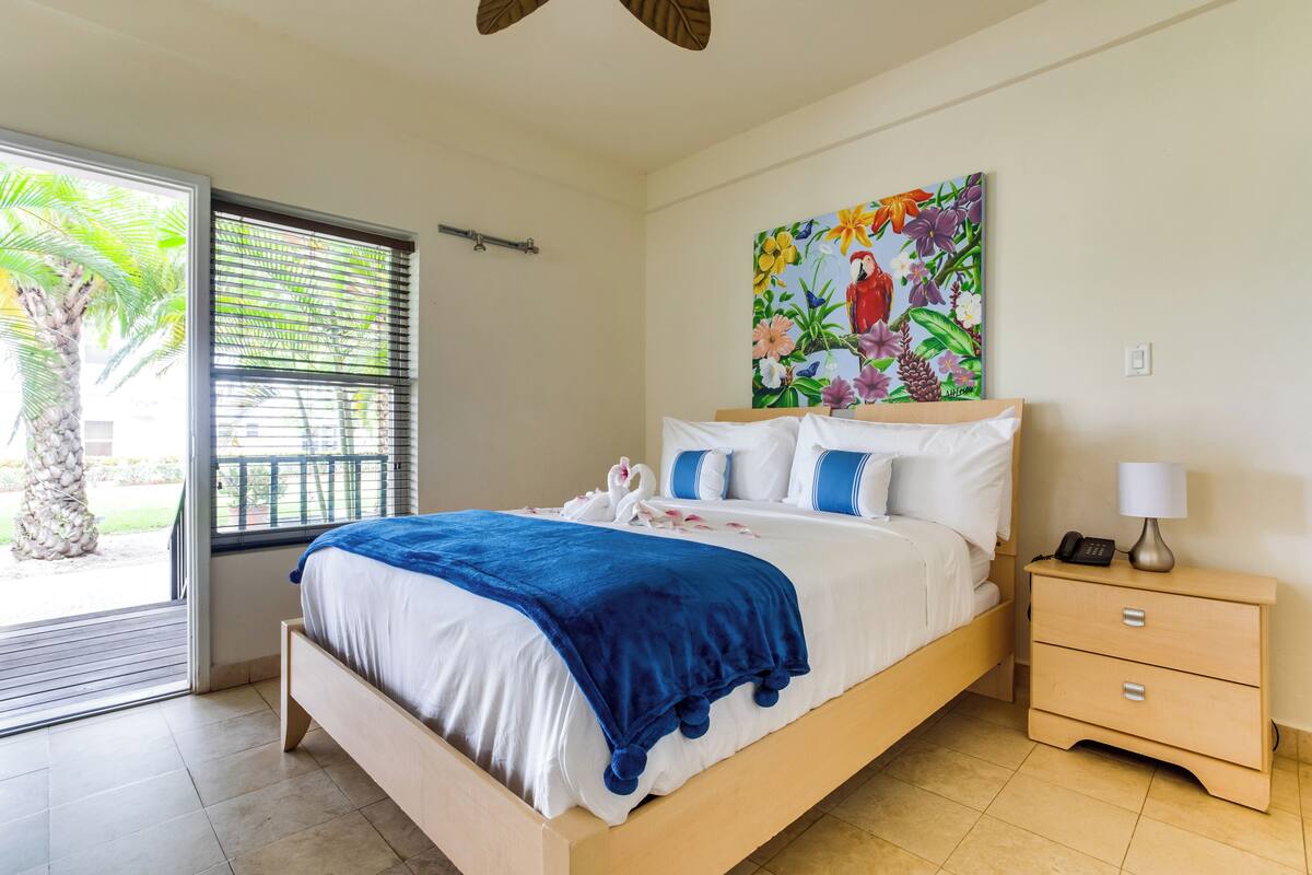 A bedroom with a bed, a nightstand, a painting, and a view of a palm tree through the window.