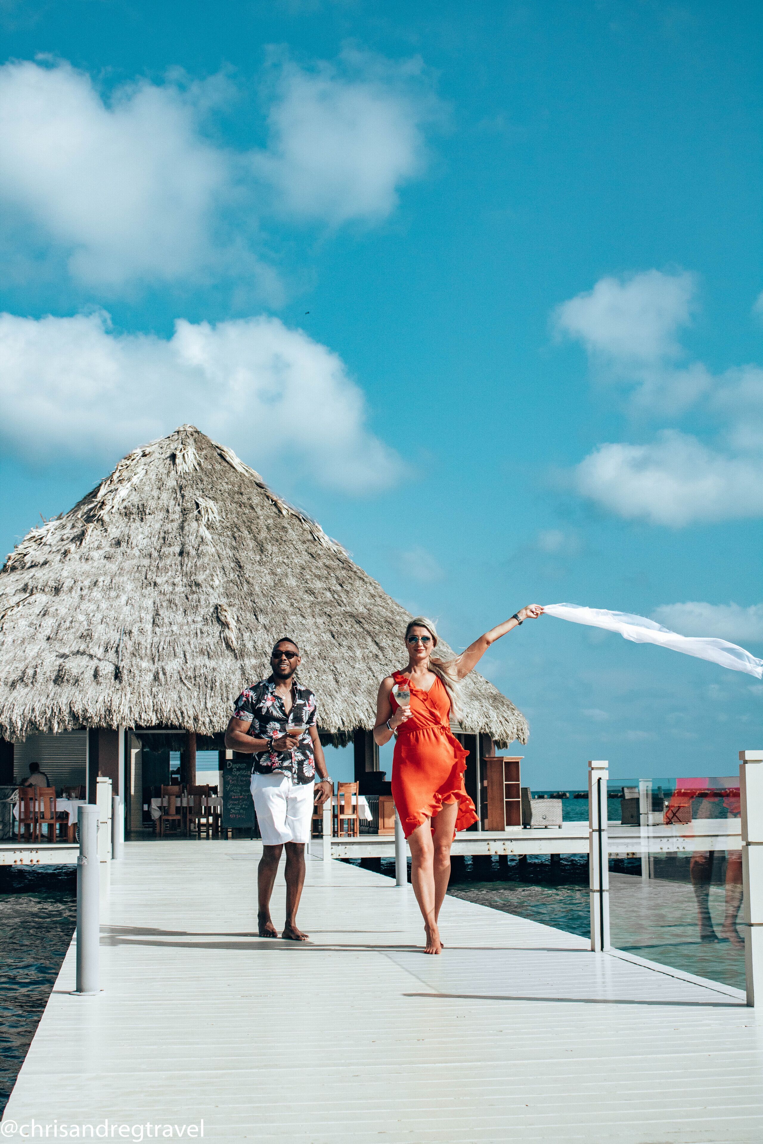 A couple walking on a wooden pier with a thatched-roof building in the background.