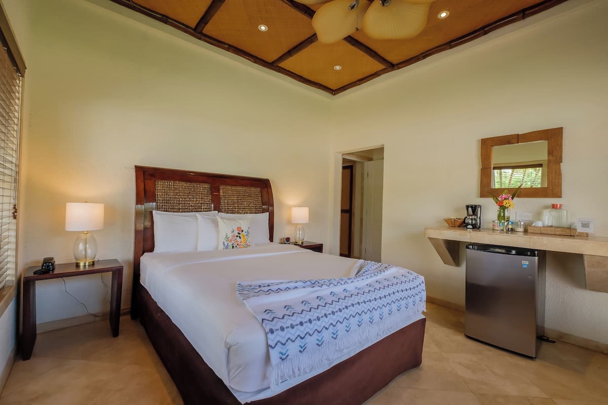 A hotel room with a large bed, bedside tables with lamps, a built-in refrigerator, and a wooden ceiling.