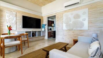 Una sala moderna con techo de madera, televisor de pantalla plana, comedor con mesa y sillas de madera, sofá, y zona de cocina con armarios y dispensador de agua.
