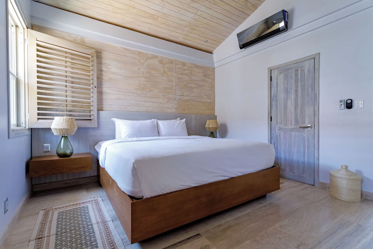 A bedroom with a wooden bed, a nightstand with lamps, a door, and a window with blinds.