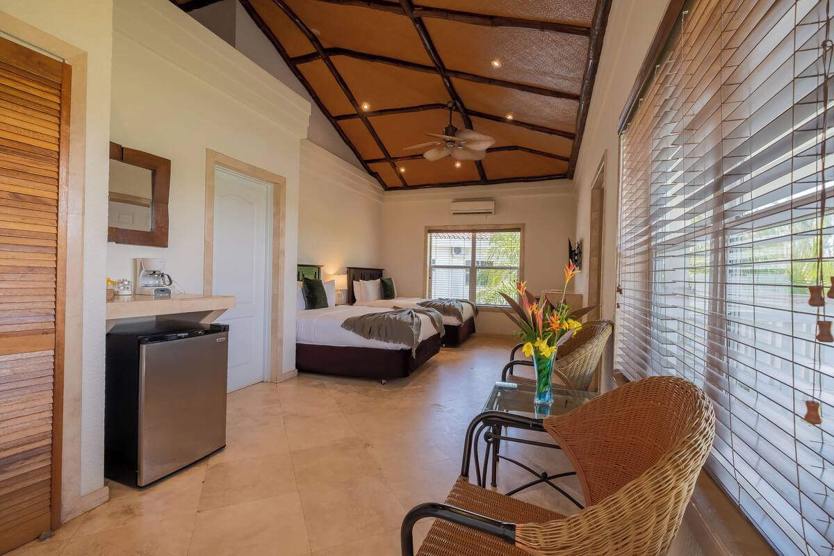 A compact hotel room with a kitchenette, a bed, a dining area with wicker furniture, and large windows with blinds.