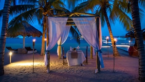 Escena de playa con mesa y sillas bajo una sombrilla, palmeras y una estructura de techo de paja.