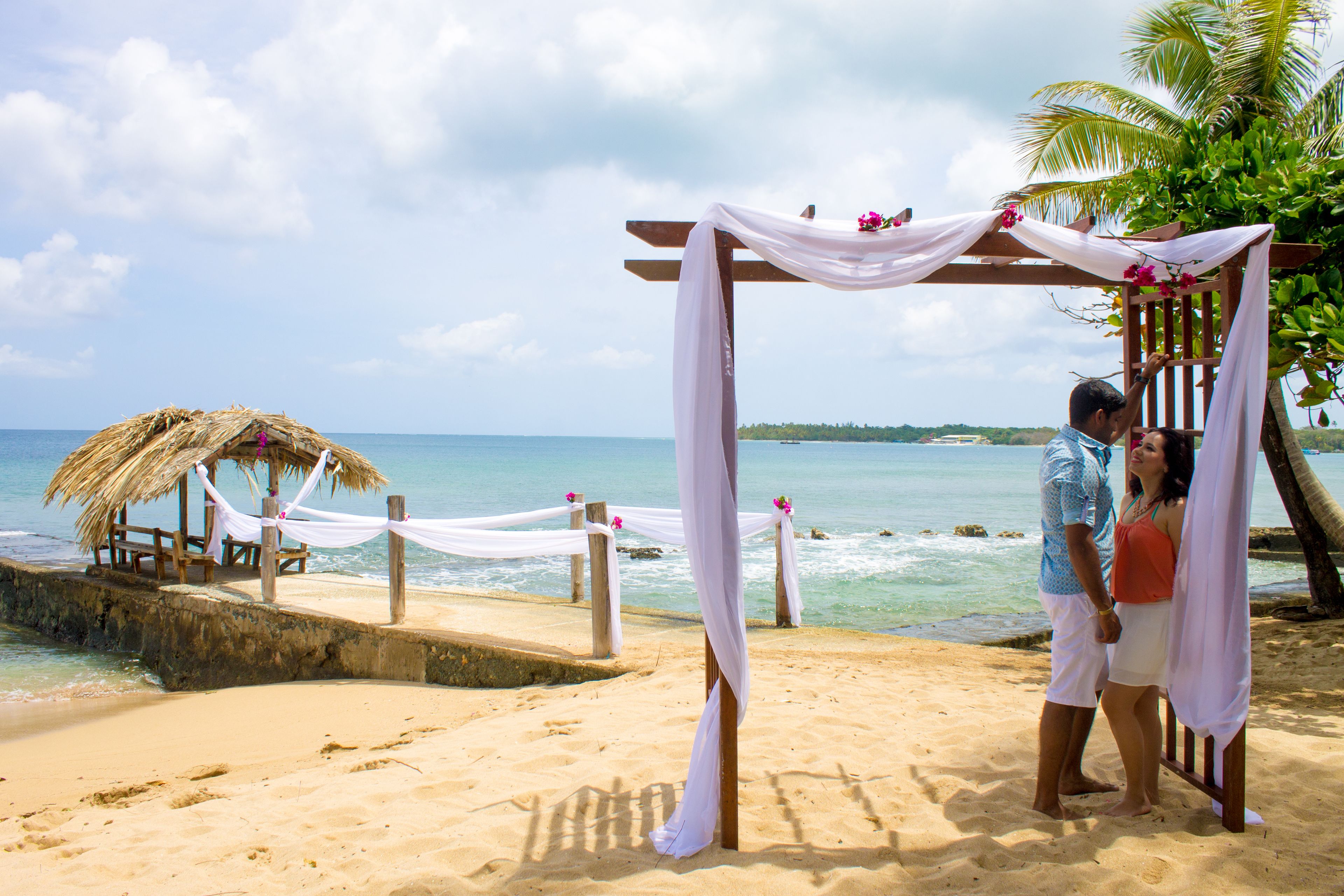 A beach wedding setup with a wooden arch and draped fabric, palm trees, and a thatched structure in the background.
