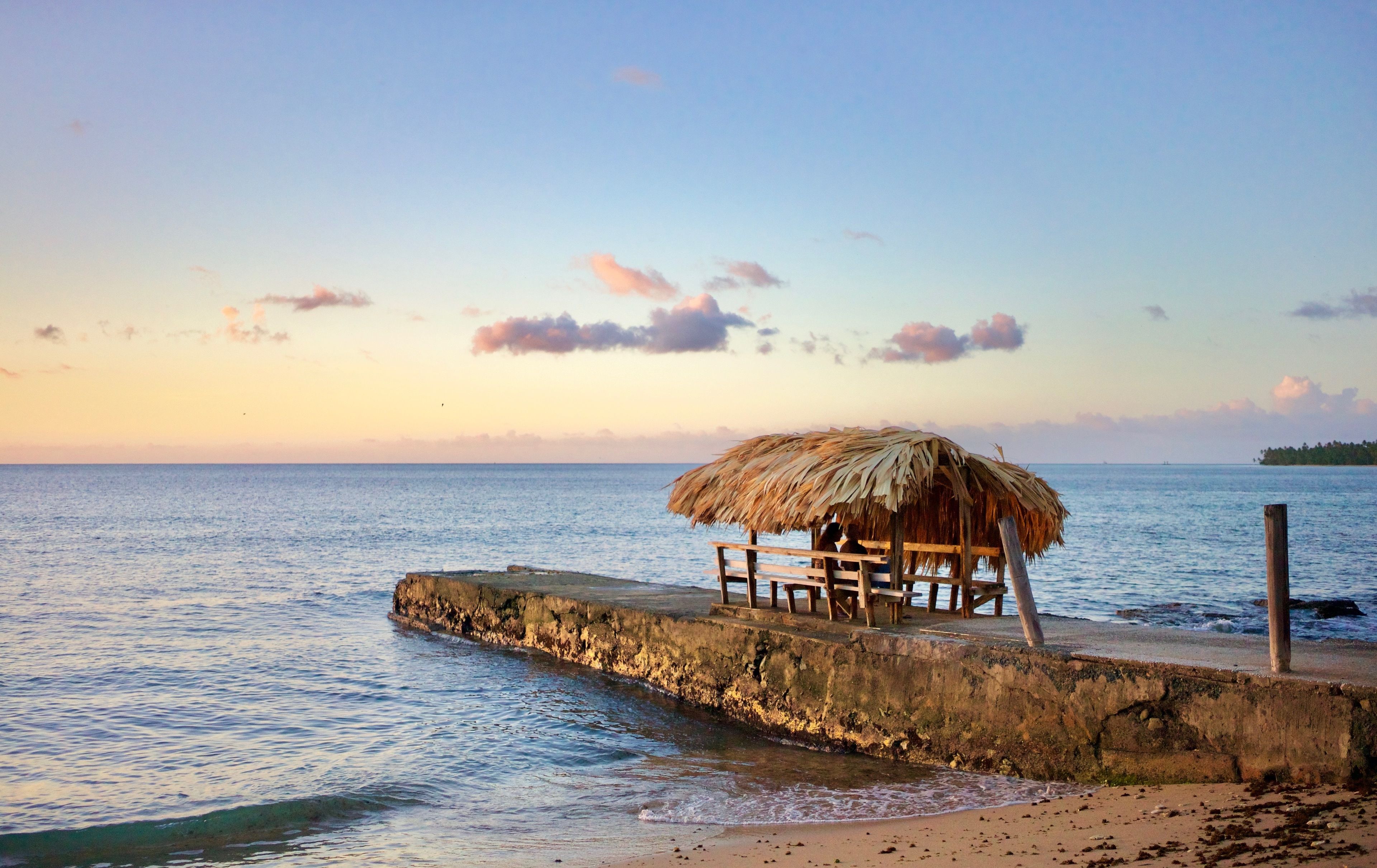 A beach with a thatched-roof hut and a wooden bench by the water.