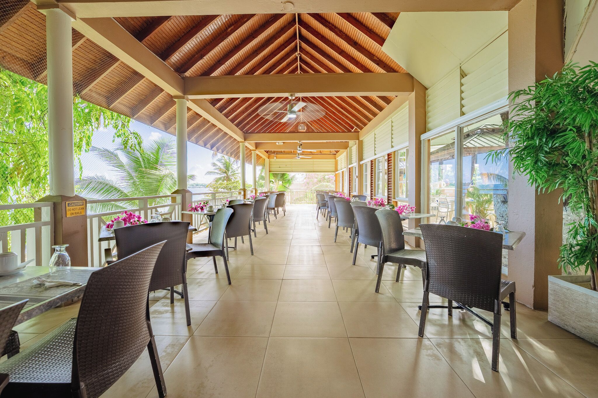 A covered outdoor dining area with tables and chairs, large windows, and a view of palm trees.