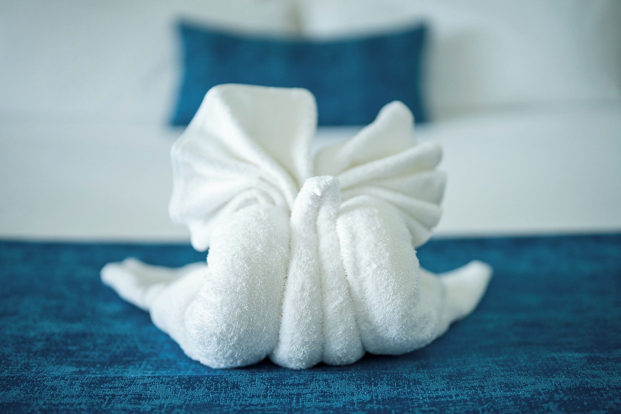A white towel folded into a bow shape on a blue textured surface, likely a bed or a table, with a blurred background suggesting a hotel room setting.