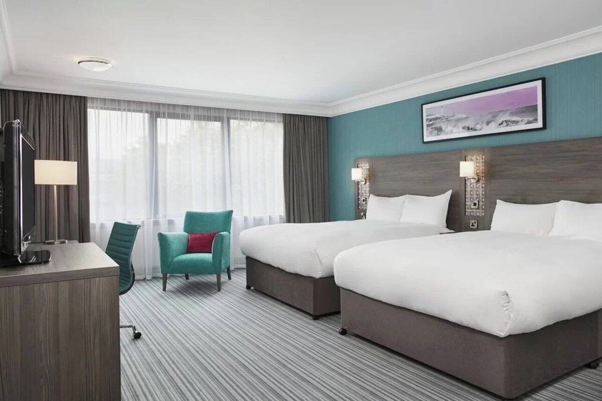 A hotel room with two beds, a desk, a chair, and a large window with curtains.