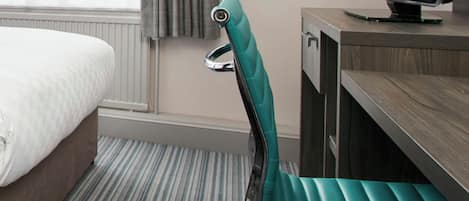 Una habitación de hotel con cama, escritorio con monitor de computadora y una silla color turquesa.
