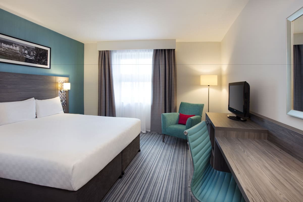A hotel room with a large bed, a desk with a television, a chair, and a window with curtains.