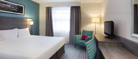 A hotel room with a large bed, a desk with a television, a chair, and a window with curtains.