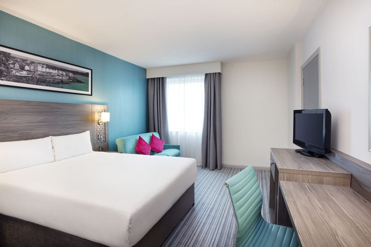 A hotel room with a large bed, a desk with a television, a chair, and a window with curtains.